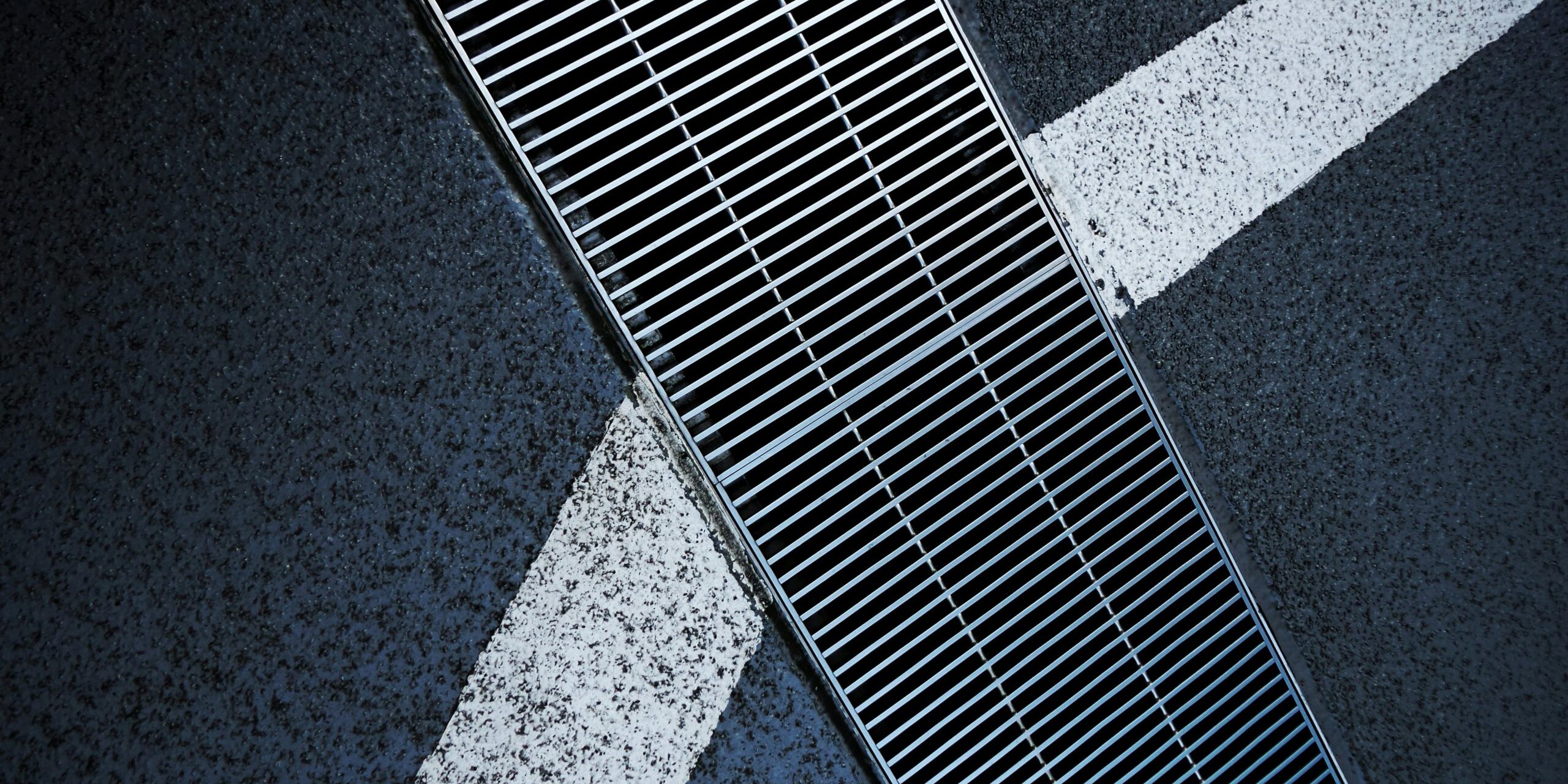 Metallic street grate intersecting white road markings on a textured asphalt surface.